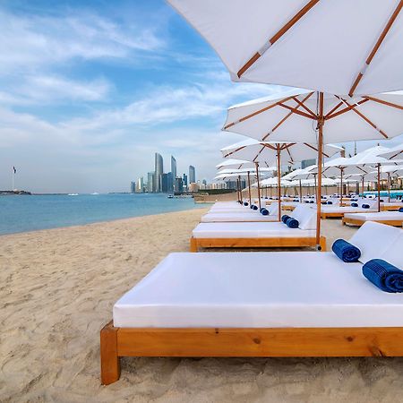 Radisson Blu Hotel & Resort, Abu Dhabi Corniche Esterno foto The photo depicts a serene beach setting with several white lounge beds arranged neatly on the sandy shore. Each bed is paired with a white umbrella providing shade. In the background, a skyline of modern buildings can be seen along the coast, hintin