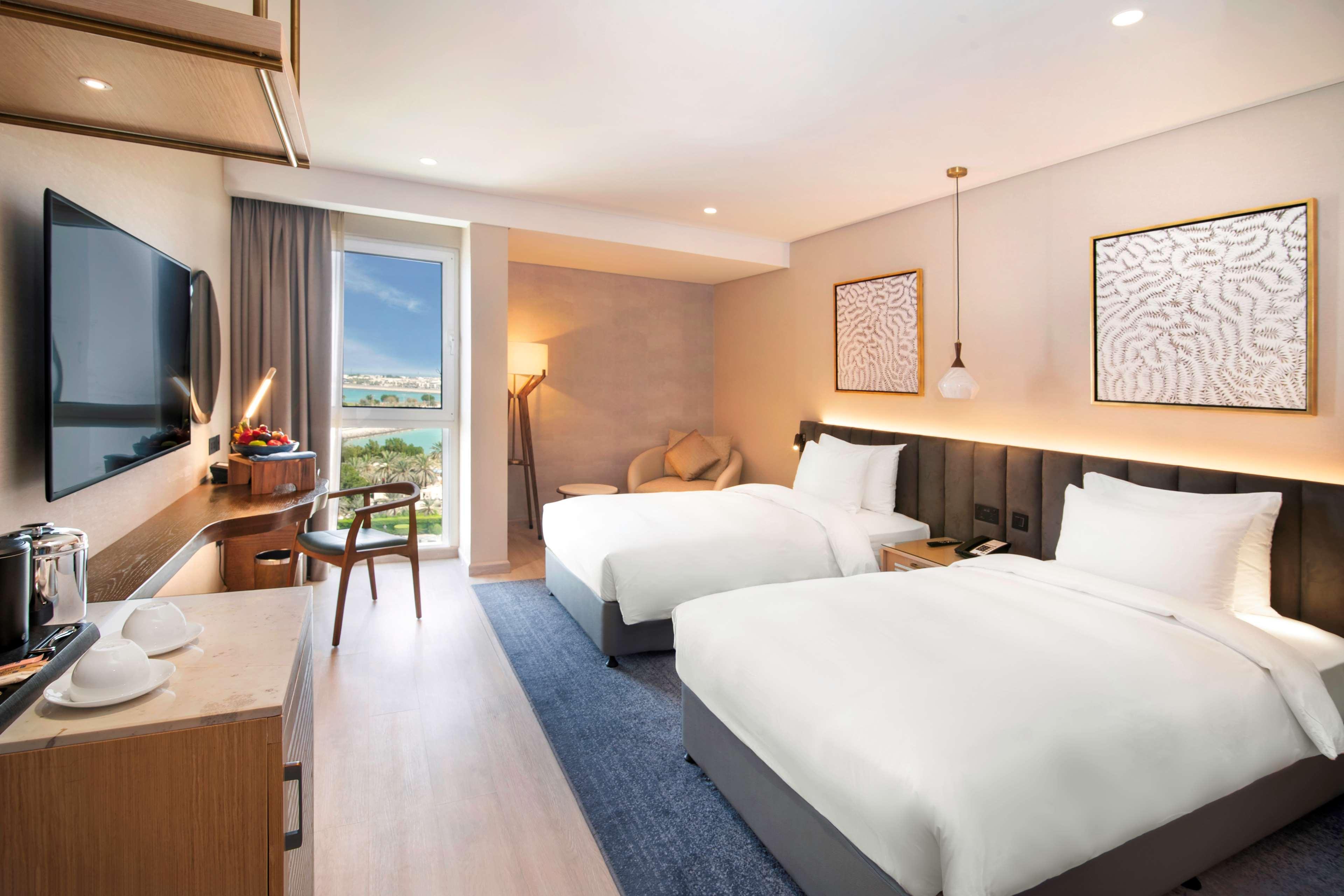 Radisson Blu Hotel & Resort, Abu Dhabi Corniche Esterno foto The photo shows a modern hotel room featuring two beds with white bedding. There is a stylish desk and chair positioned near a large window that allows natural light to fill the space. The room has a contemporary design with muted colors and decorati