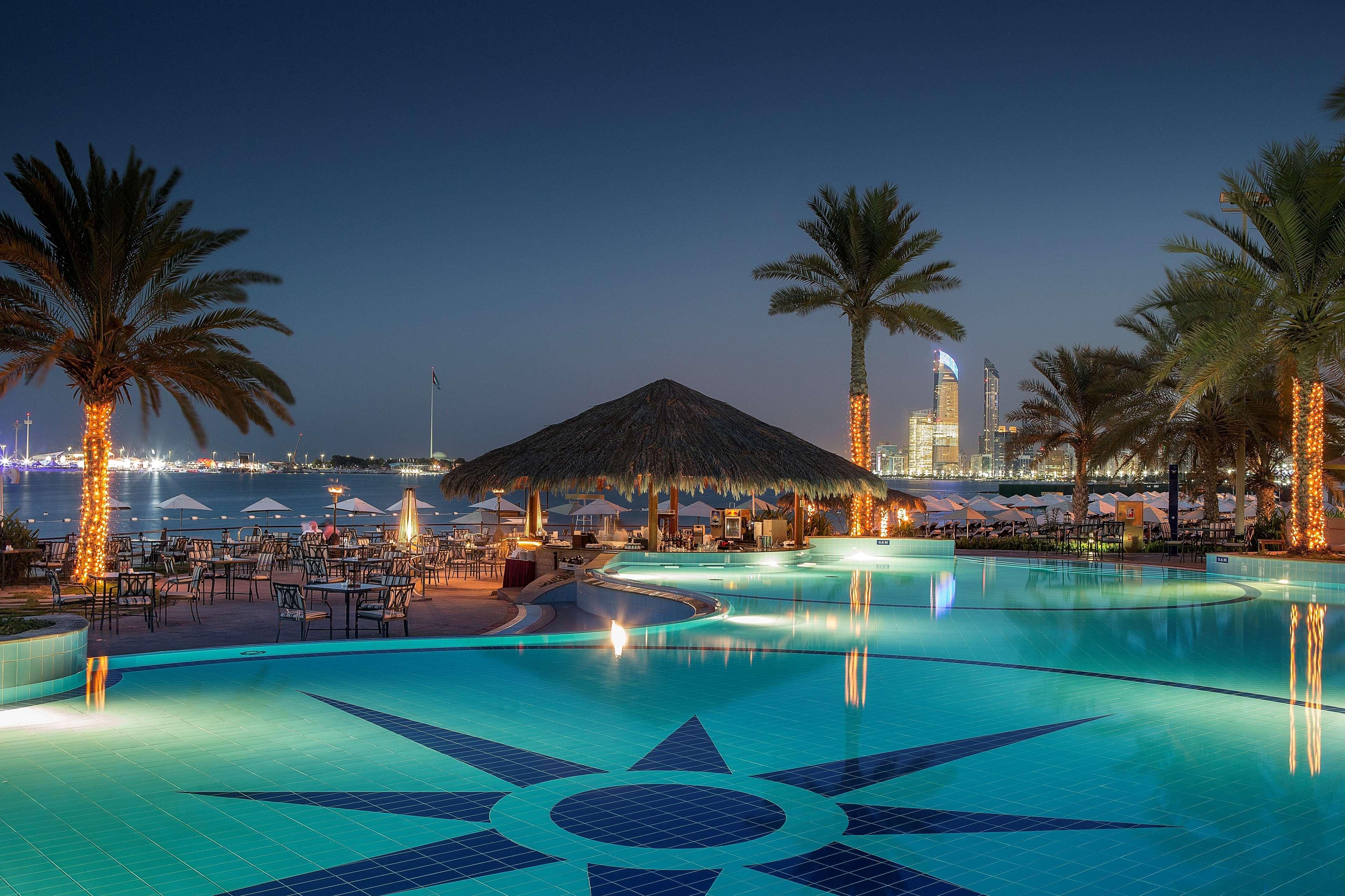 Radisson Blu Hotel & Resort, Abu Dhabi Corniche Esterno foto The photo shows a picturesque outdoor pool area at night. The pool features a decorative tile pattern resembling a star at its center. Surrounding the pool are tall palm trees, some illuminated by soft lighting. In the background, there is a beachsid