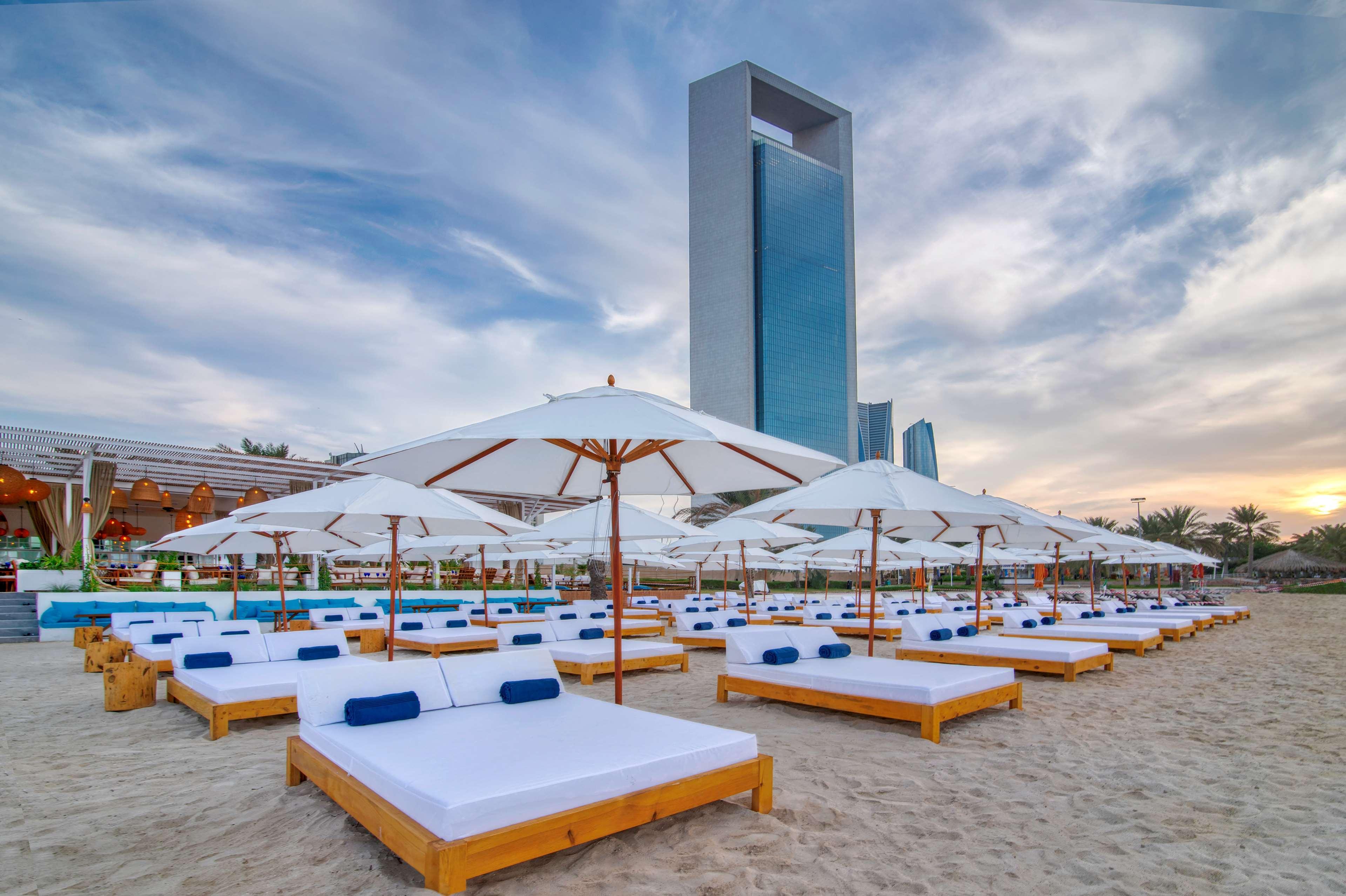 Radisson Blu Hotel & Resort, Abu Dhabi Corniche Esterno foto The photo depicts a beach scene with a number of wooden sunbeds arranged on the sandy shore. Each sunbed is equipped with white cushions and blue pillows. Large white umbrellas are positioned above the sunbeds, providing shade. In the background, the