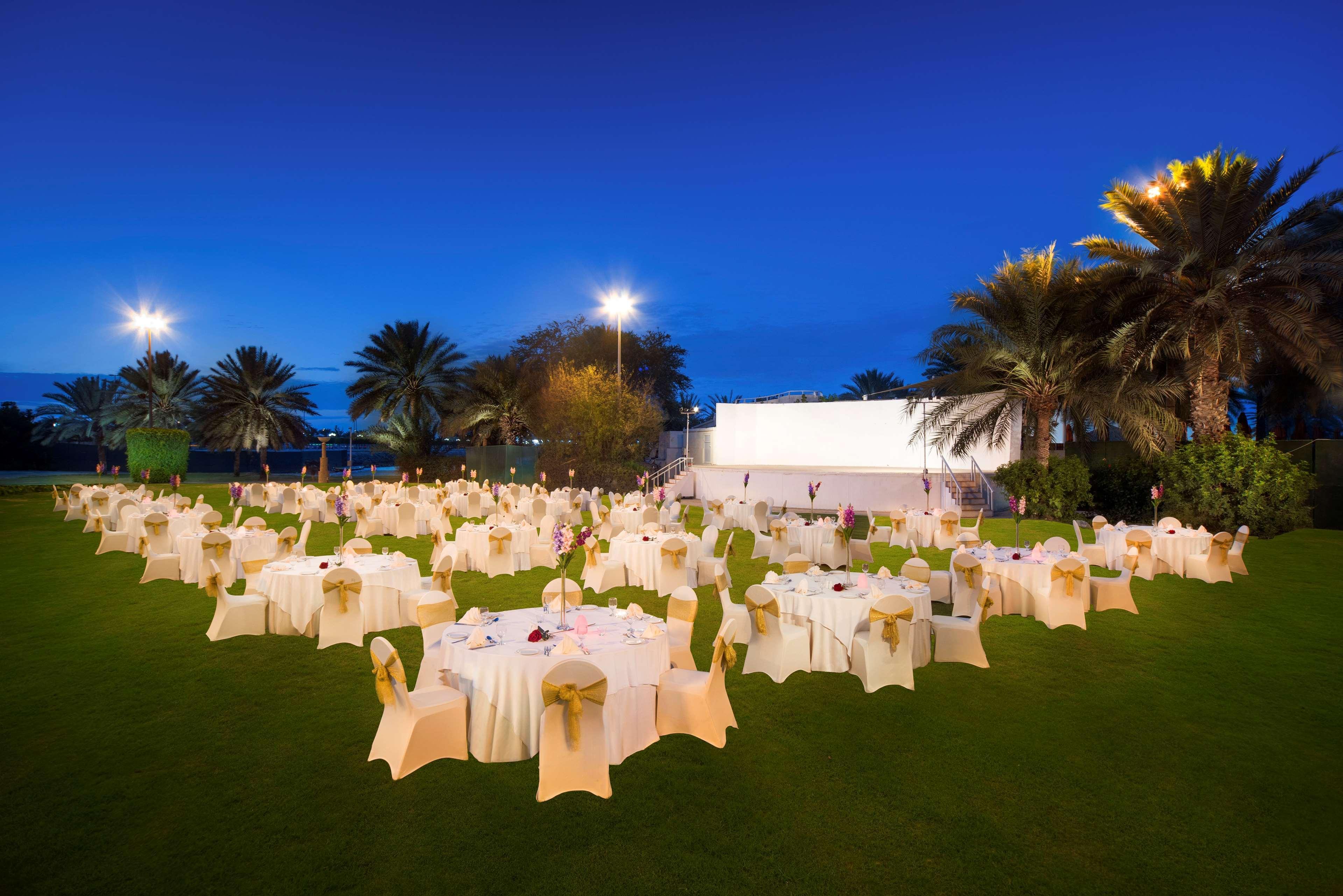 Radisson Blu Hotel & Resort, Abu Dhabi Corniche Esterno foto The photo shows an outdoor venue set up for an event at dusk. There are numerous round tables draped in white tablecloths, each adorned with pale gold sashes on the chairs. The setting is landscaped with lush greenery and palm trees. In the backgroun