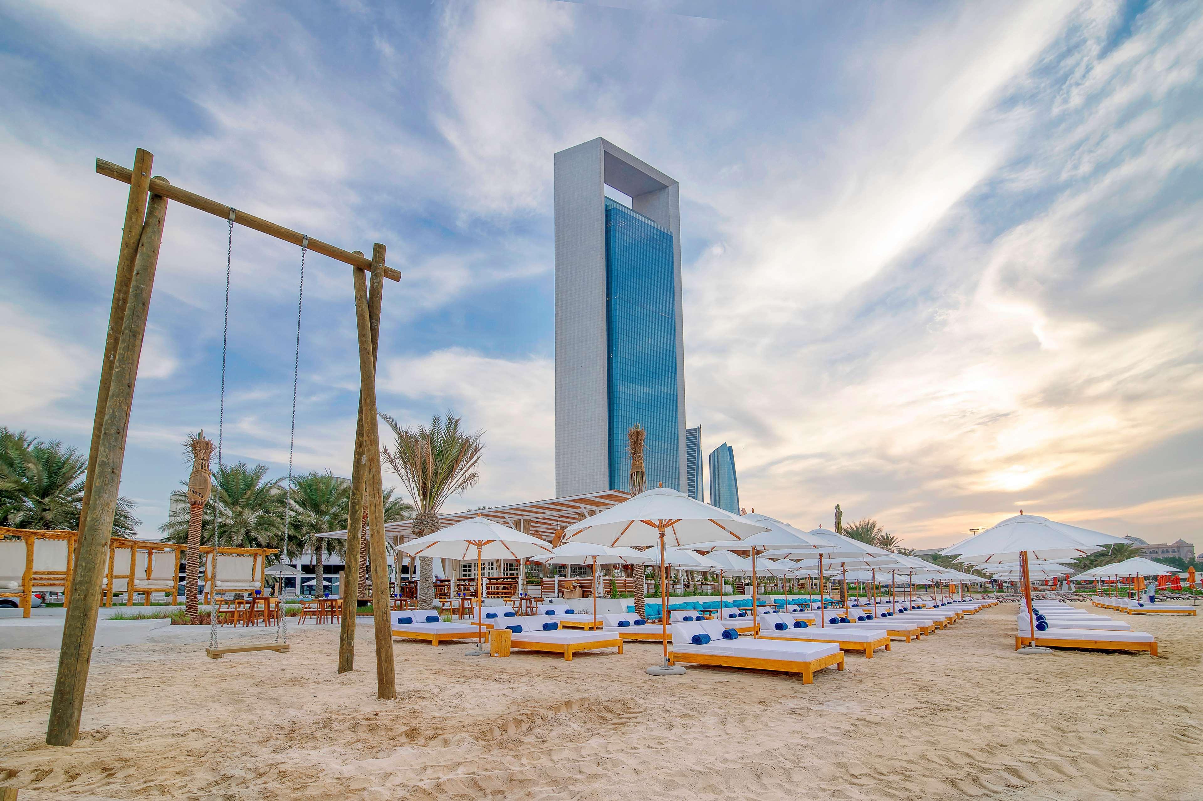 Radisson Blu Hotel & Resort, Abu Dhabi Corniche Esterno foto The photo shows a beach scene with several sun loungers arranged under white umbrellas, set against a backdrop of a tall, modern building. There are palm trees in the area, providing a tropical ambiance. On one side, there's a wooden swing set, addin