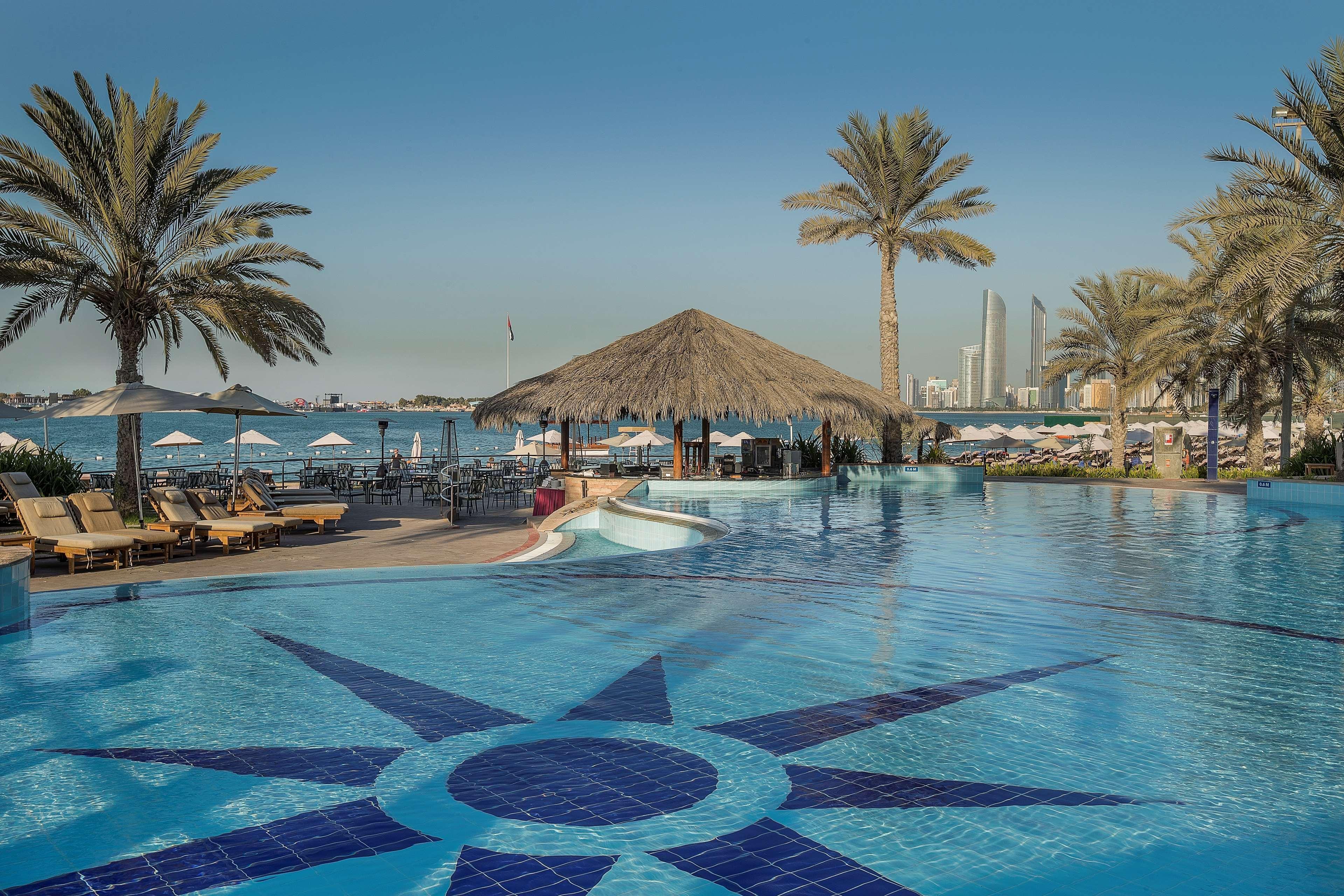 Radisson Blu Hotel & Resort, Abu Dhabi Corniche Esterno foto The photo shows a panoramic view of a luxurious resort pool area. In the foreground, there is a large blue swimming pool with a decorative star pattern on the bottom. Surrounding the pool are several lounge chairs, and there are palm trees providing 