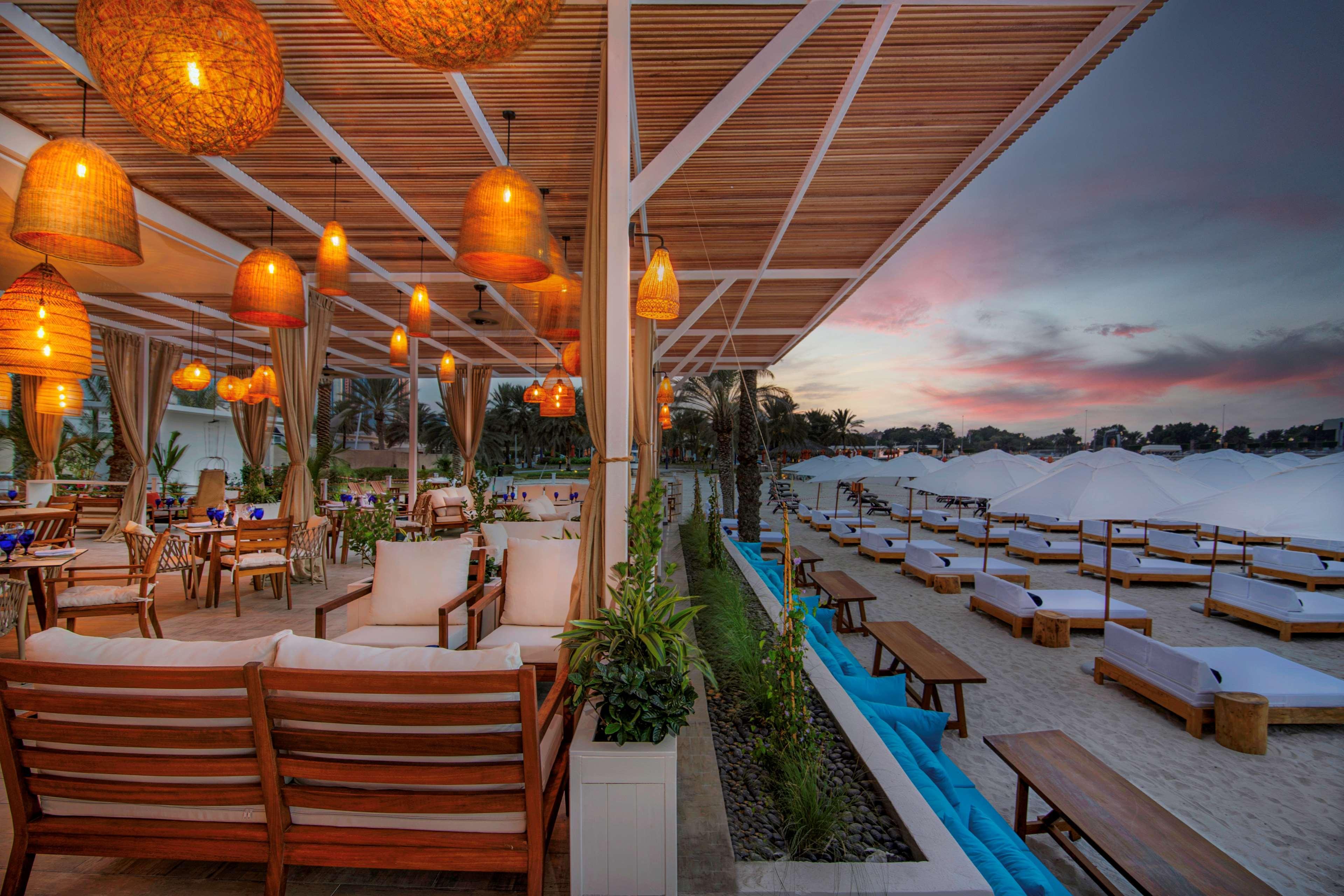 Radisson Blu Hotel & Resort, Abu Dhabi Corniche Esterno foto The photo depicts a stylish beachside lounge or restaurant. In the foreground, there are modern wooden chairs and tables arranged comfortably for guests, along with plush seating options. The space is decorated with hanging pendant lights, creating a