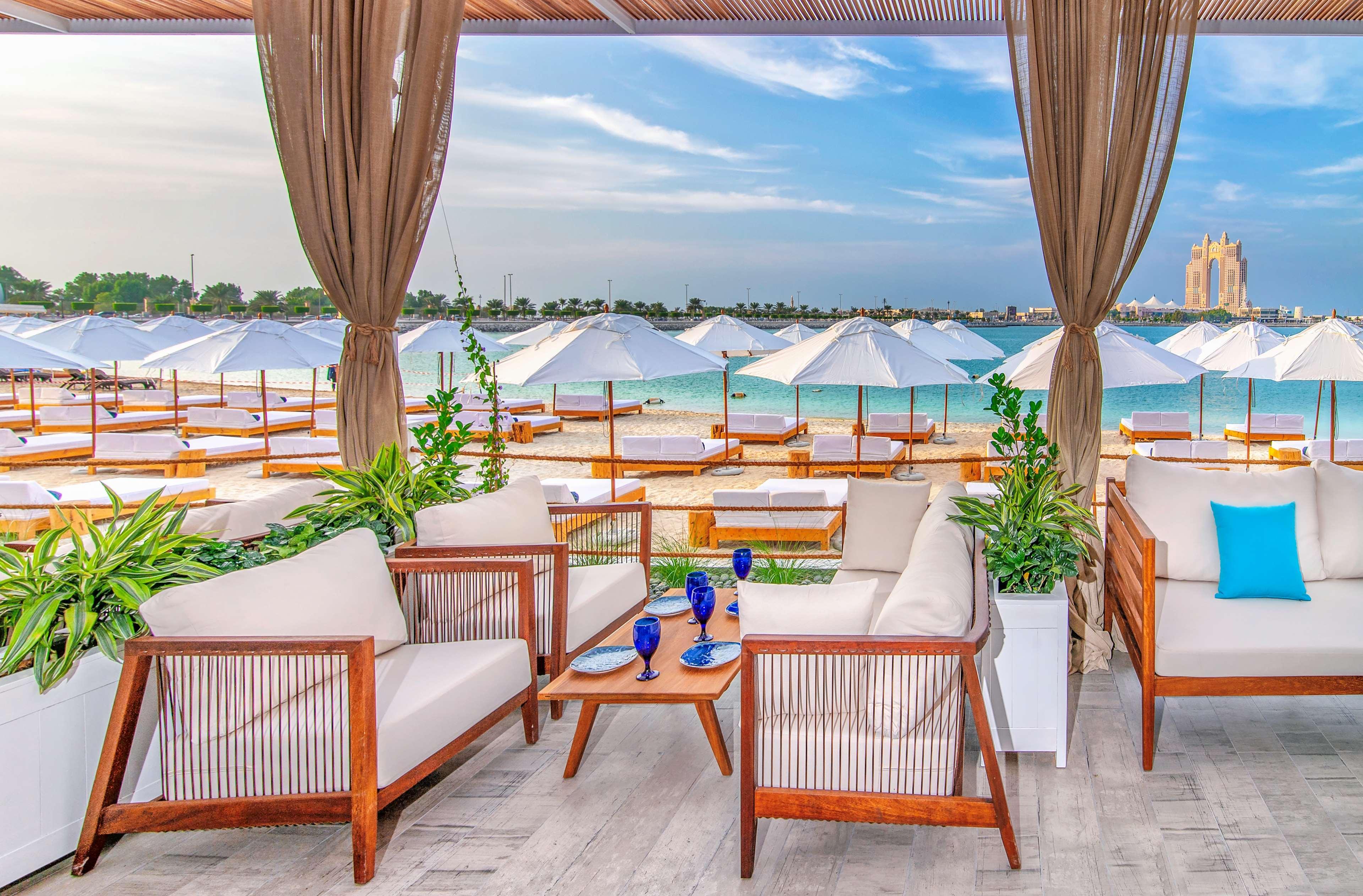 Radisson Blu Hotel & Resort, Abu Dhabi Corniche Esterno foto The photo shows a stylish outdoor seating area at a beach or resort. There are comfortable lounge sofas arranged around a wooden coffee table, accompanied by decorative plants. In the background, a sandy beach is visible, lined with white umbrellas a