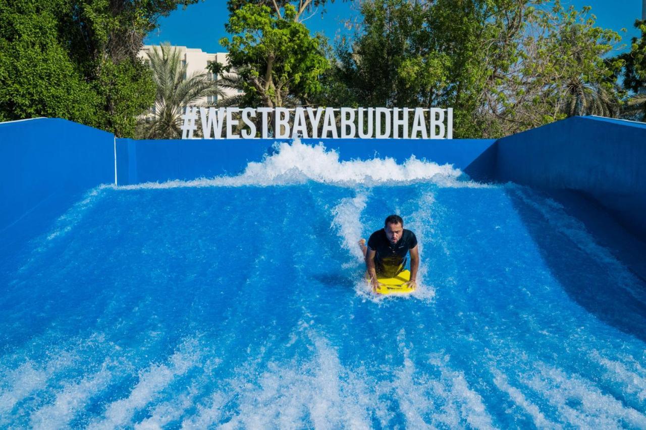 Radisson Blu Hotel & Resort, Abu Dhabi Corniche Esterno foto The photo shows a person riding a bodyboard on a wave simulator, creating artificial waves. The background features a bright blue wall with the hashtag #WESTBAYABUDHABI displayed prominently. Surrounding the wave pool, there are trees and greenery, s
