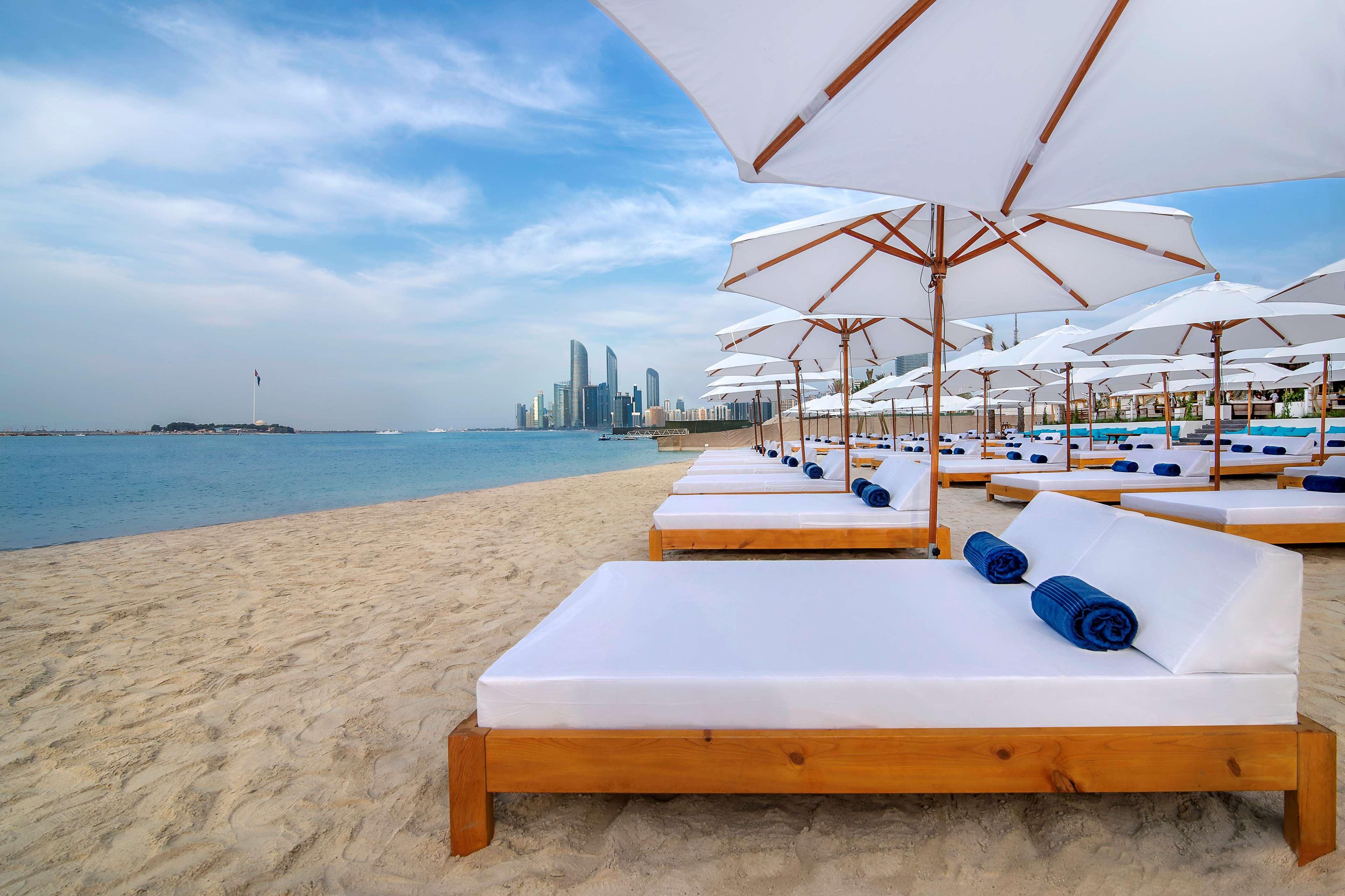 Radisson Blu Hotel & Resort, Abu Dhabi Corniche Esterno foto The photo depicts a serene beach setting with several white lounge beds arranged neatly on the sandy shore. Each bed is paired with a white umbrella providing shade. In the background, a skyline of modern buildings can be seen along the coast, hintin