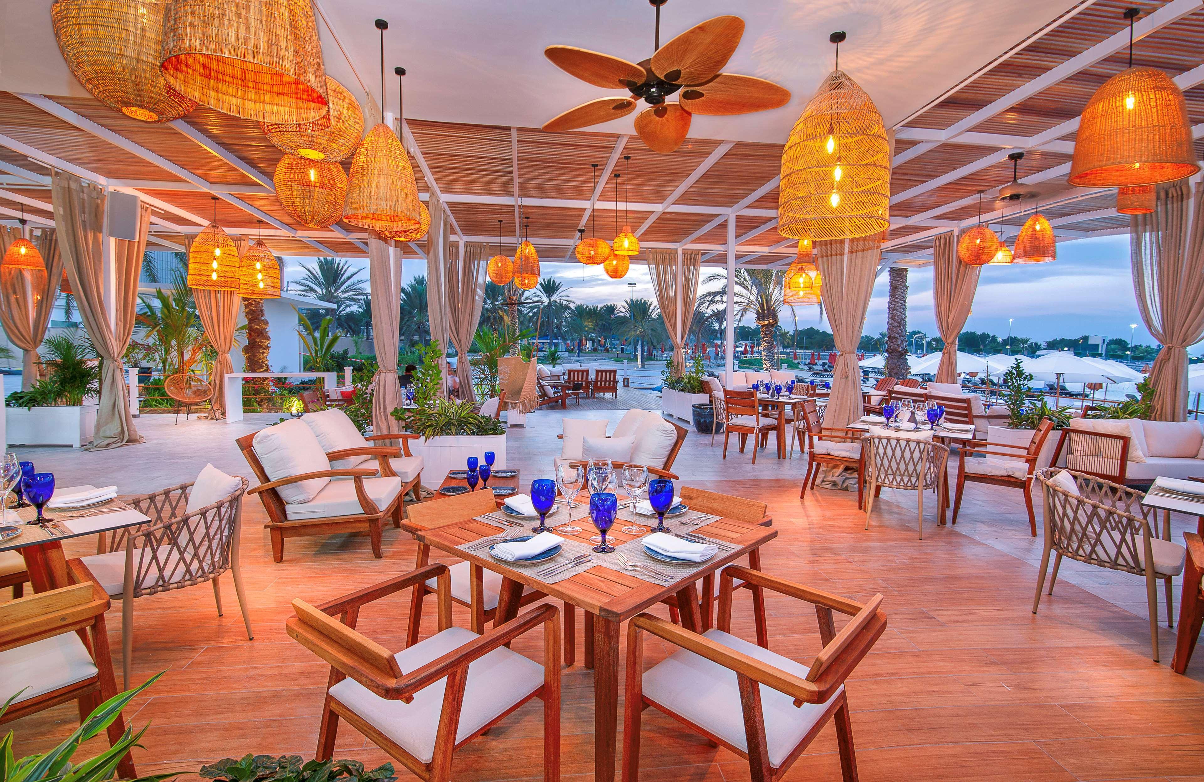 Radisson Blu Hotel & Resort, Abu Dhabi Corniche Esterno foto The photo depicts an elegant dining area, likely in a restaurant. The space features wooden tables and comfortable chairs arranged neatly. There are multiple blue glasses set on the tables, suggesting a readiness for dining. The lighting is warm, pro