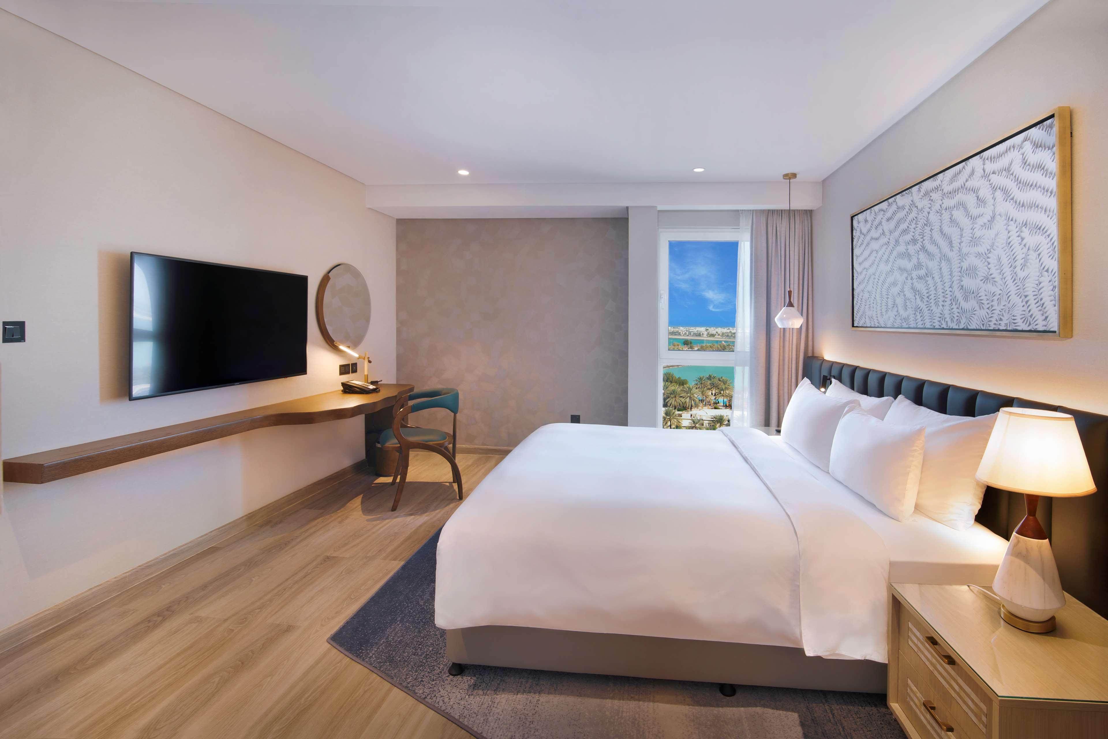 Radisson Blu Hotel & Resort, Abu Dhabi Corniche Esterno foto The photo shows a modern hotel room with a clean and minimalistic design. It features a large bed with white bedding, complemented by bedside tables on either side with lamps. There's a wall-mounted television opposite the bed, and a desk with a chai