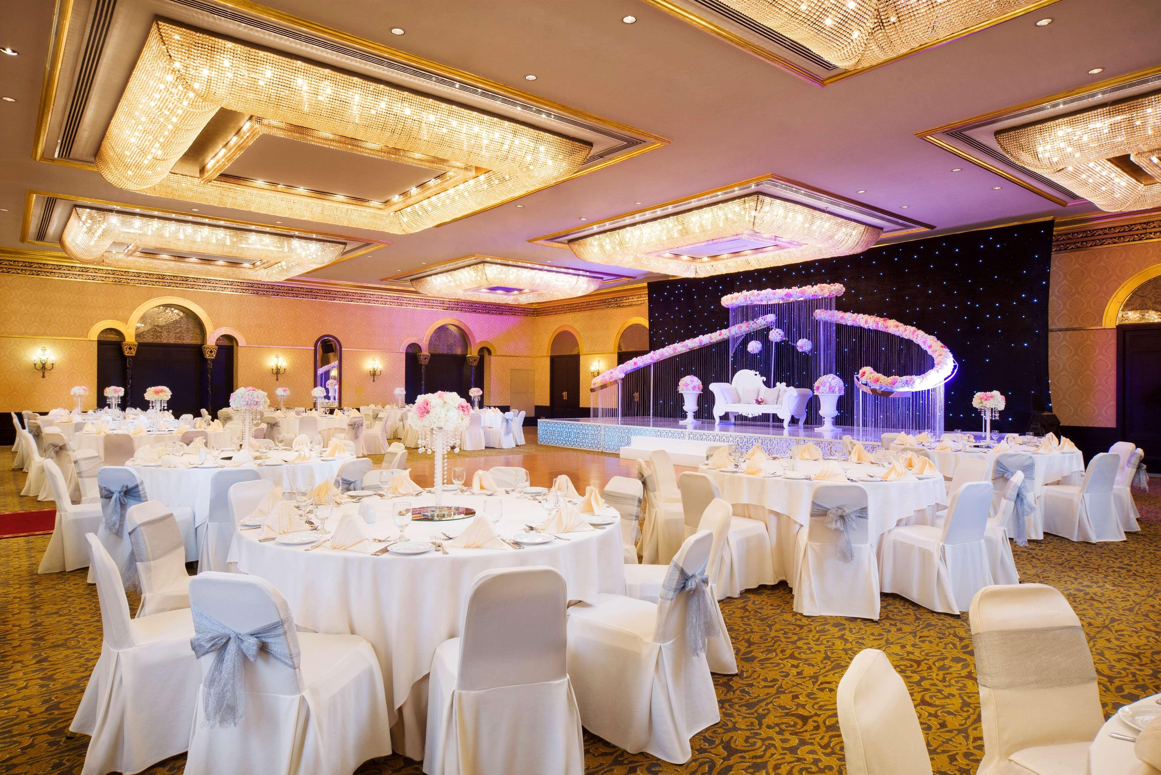Radisson Blu Hotel & Resort, Abu Dhabi Corniche Esterno foto The image shows an elegantly decorated banquet or event hall. There are several round tables covered with white tablecloths, each set with plates, cutlery, and folded napkins. The chairs are covered in white fabric with light blue sashes tied around 