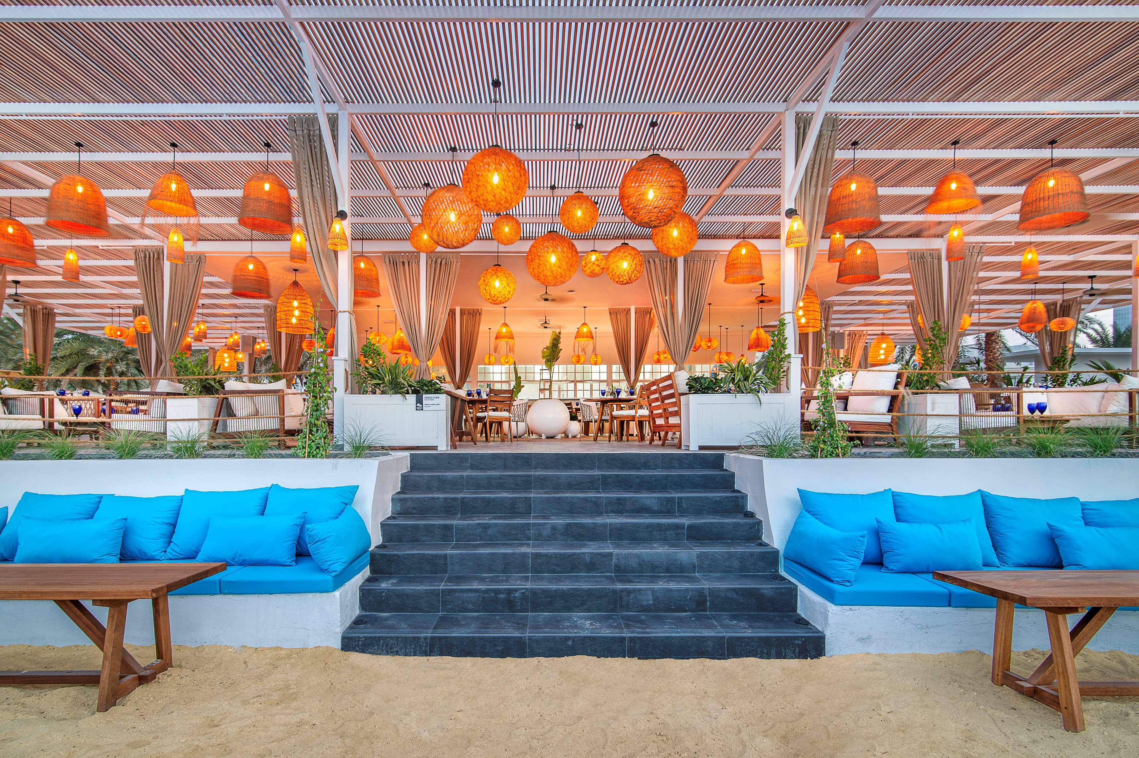 Radisson Blu Hotel & Resort, Abu Dhabi Corniche Esterno foto The photo shows a modern, stylish restaurant or lounge area that appears to be located near a beach. The setting features a spacious outdoor dining space with a roof supported by beams. The interior is decorated with warm lighting lamps, which are ha
