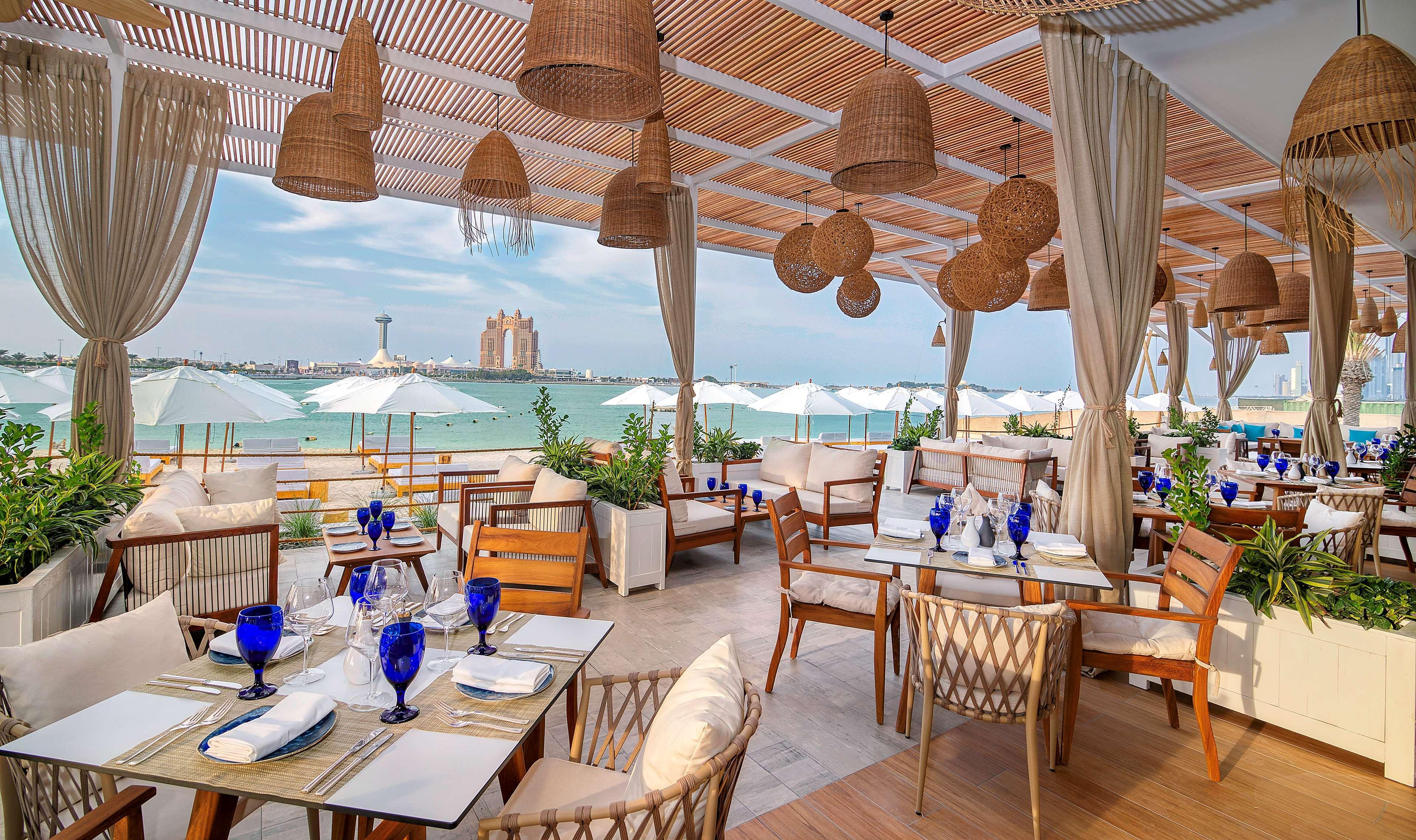 Radisson Blu Hotel & Resort, Abu Dhabi Corniche Esterno foto The photo shows a beautifully designed restaurant setting located by the beach. The interior features wooden furniture, with tables set for dining. Each table is adorned with blue glasses and neatly arranged place settings. The decor includes hanging