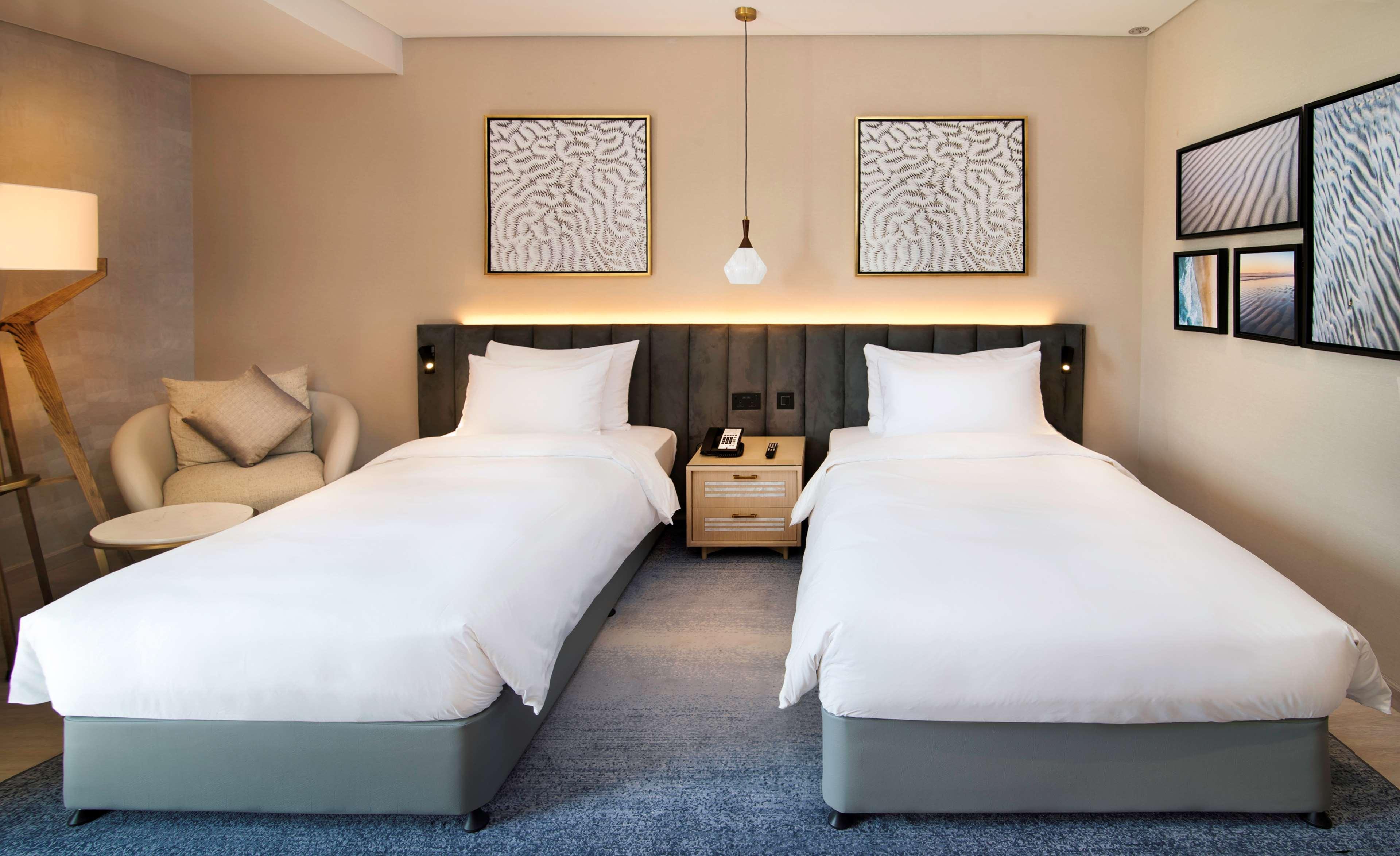 Radisson Blu Hotel & Resort, Abu Dhabi Corniche Esterno foto The photo shows a modern hotel room with two single beds, each dressed in white bedding. The beds are positioned against a headboard with a gray color scheme. Above each bed, there are two framed artworks on the wall. A pendant light hangs from the c