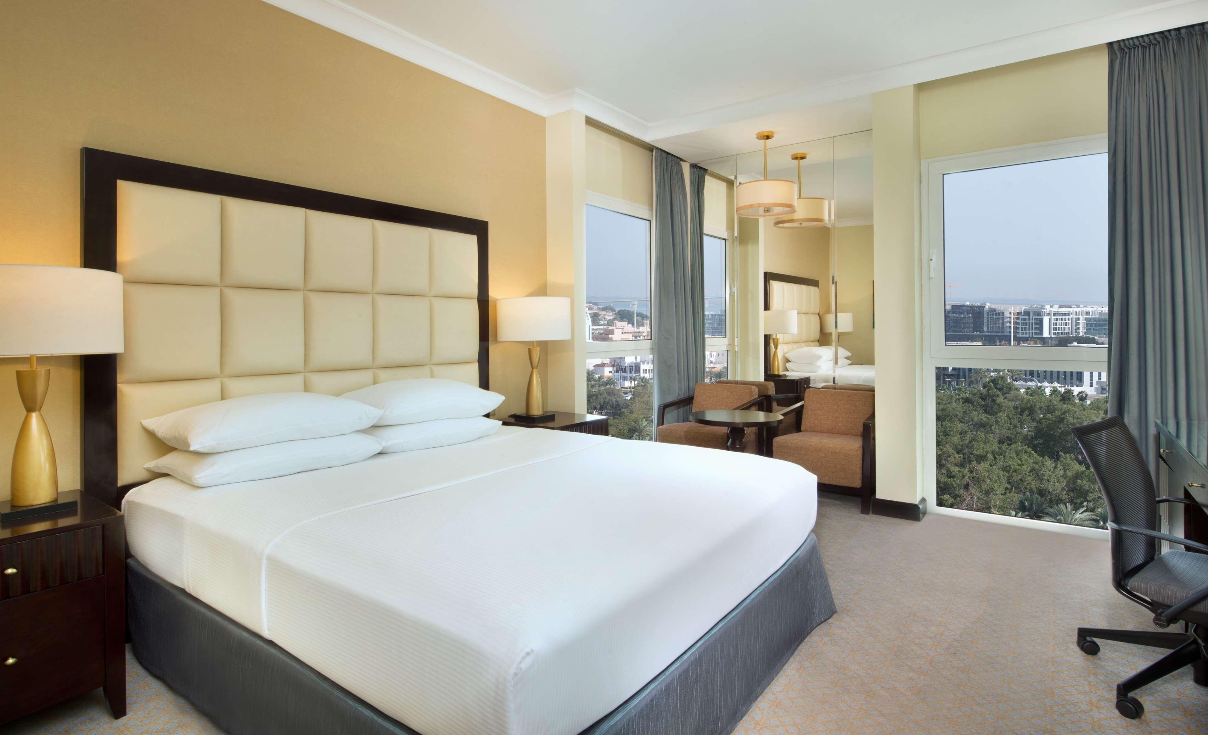Radisson Blu Hotel & Resort, Abu Dhabi Corniche Esterno foto The photo shows a modern hotel room featuring a large bed with white linens and a gray bed skirt. The headboard is upholstered in a light-colored fabric. There are bedside lamps on either side of the bed. In the background, there are windows offering
