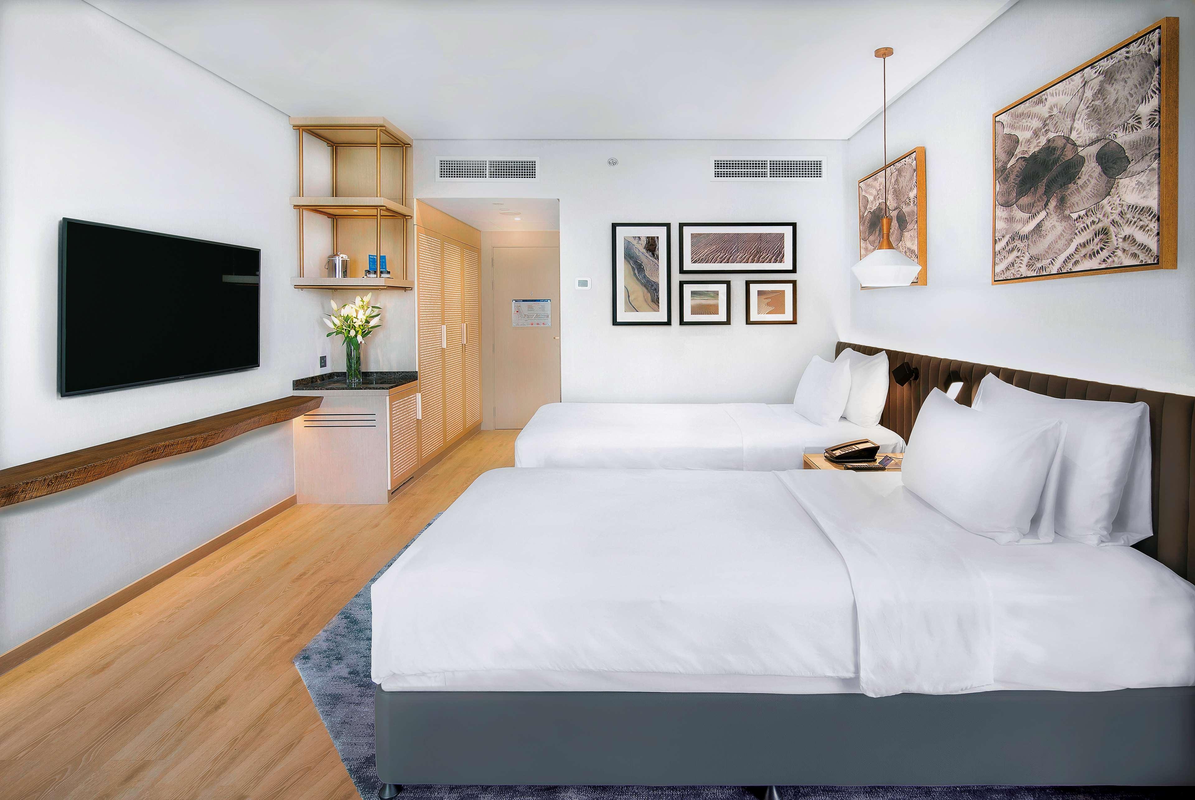 Radisson Blu Hotel & Resort, Abu Dhabi Corniche Esterno foto The photo shows a modern hotel room featuring two beds with white bedding. There's a flat-screen TV mounted on the wall opposite the beds. The room has light-colored wooden flooring and a large window, allowing natural light to enter. A small kitchen