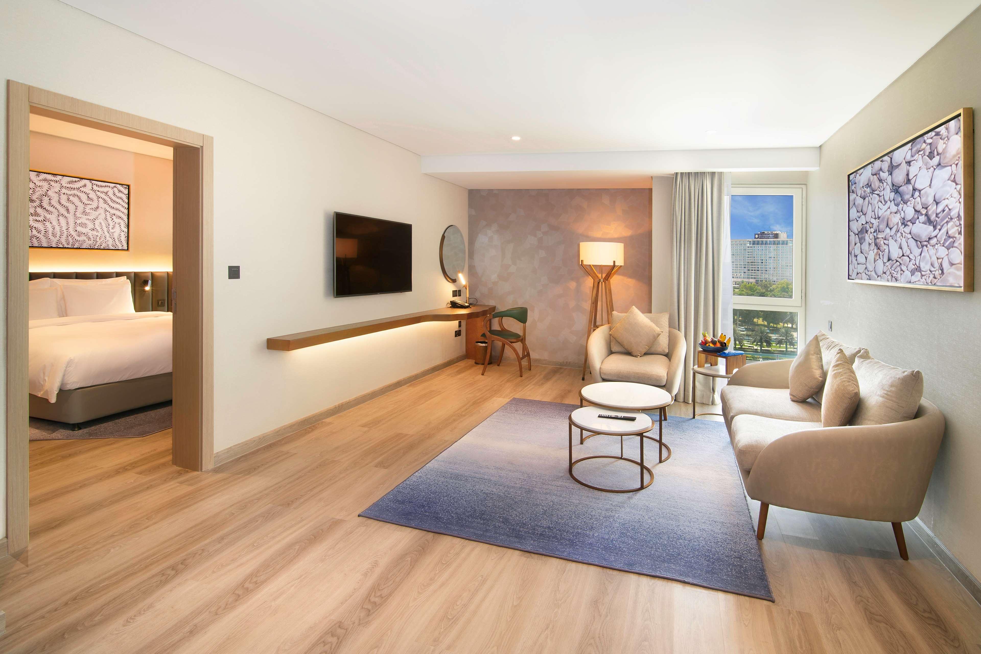Radisson Blu Hotel & Resort, Abu Dhabi Corniche Esterno foto The photo shows a modern and stylish hotel suite. It features a spacious seating area with a light-colored sofa and two armchairs arranged around a small coffee table on a soft area rug. A lamp stands next to the seating area, providing additional li