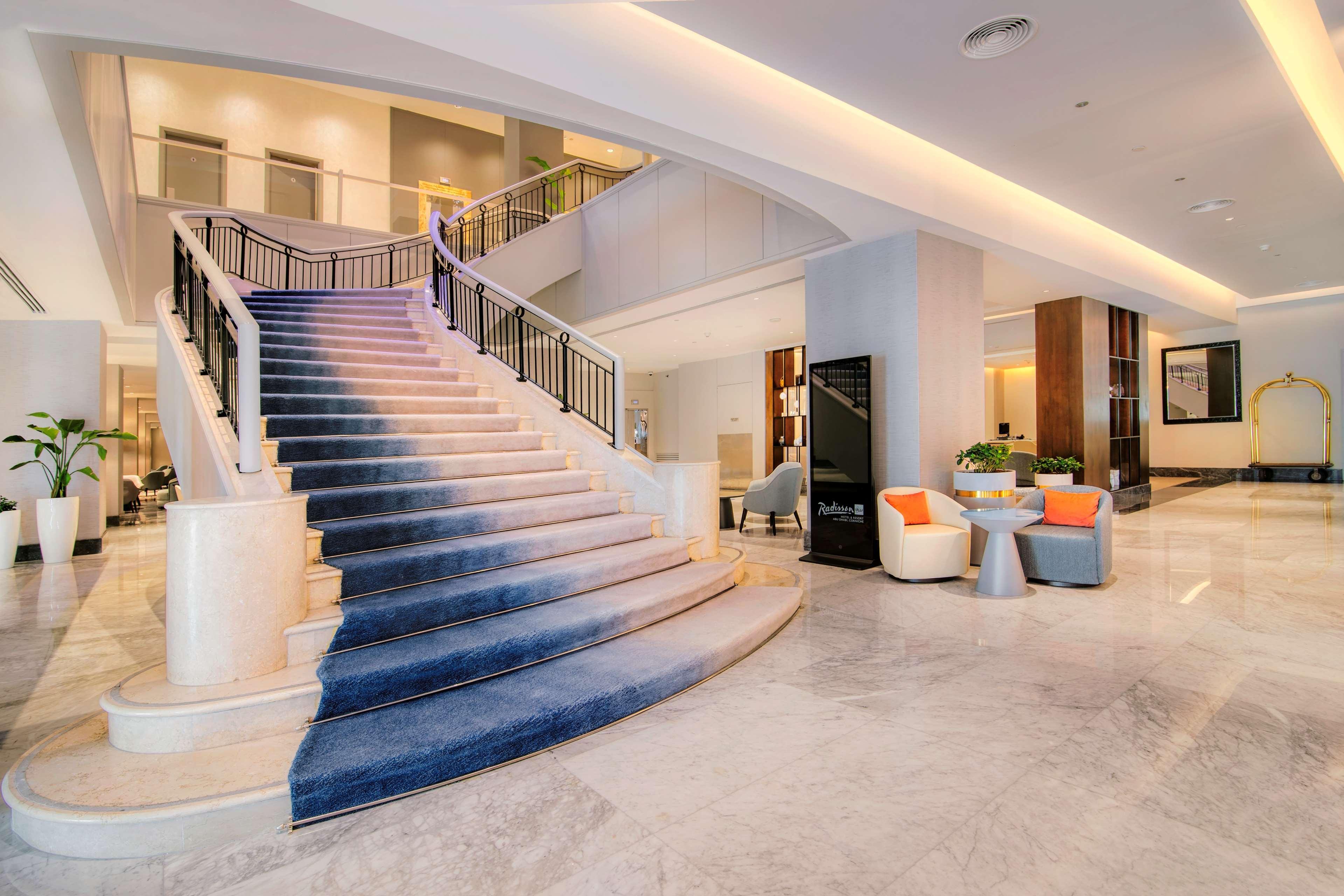 Radisson Blu Hotel & Resort, Abu Dhabi Corniche Esterno foto The photo shows a welcoming hotel lobby with a prominent staircase. The staircase features a stylish blue carpet that gradually transitions in color. The lobby is bright and spacious, with modern decor. To one side, there are seating arrangements con
