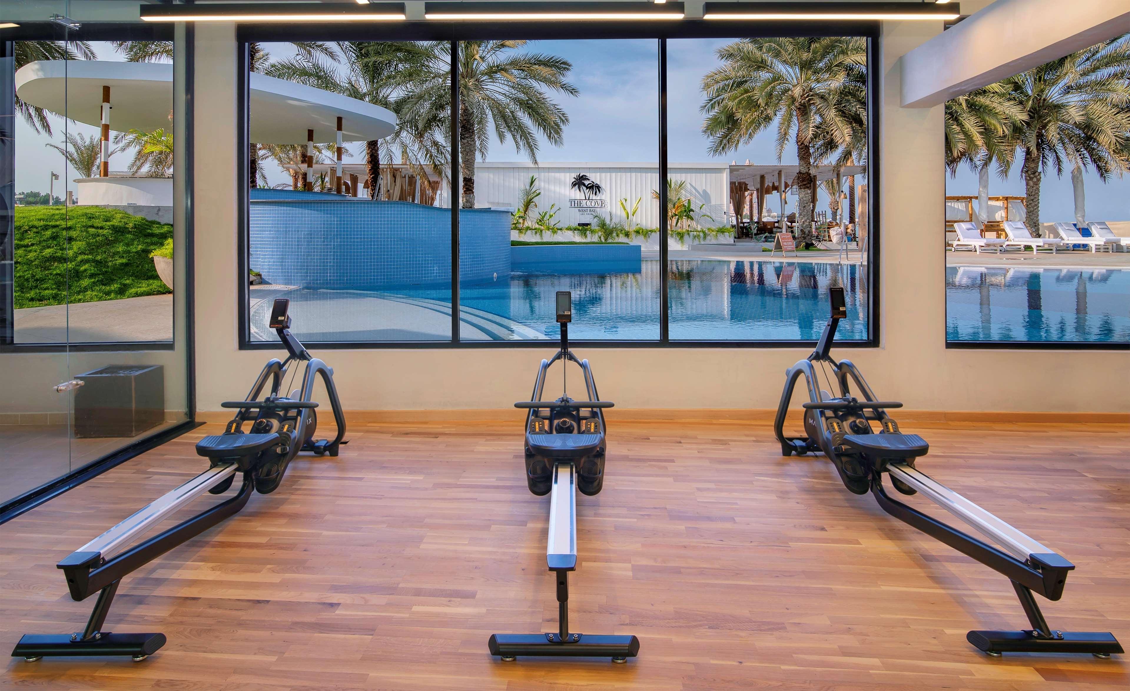 Radisson Blu Hotel & Resort, Abu Dhabi Corniche Esterno foto The photo shows a gym or fitness area featuring three rowing machines lined up on a wooden floor. In the background, there is a large window that displays an outdoor scene with palm trees and a swimming pool, creating a bright and inviting atmosphere