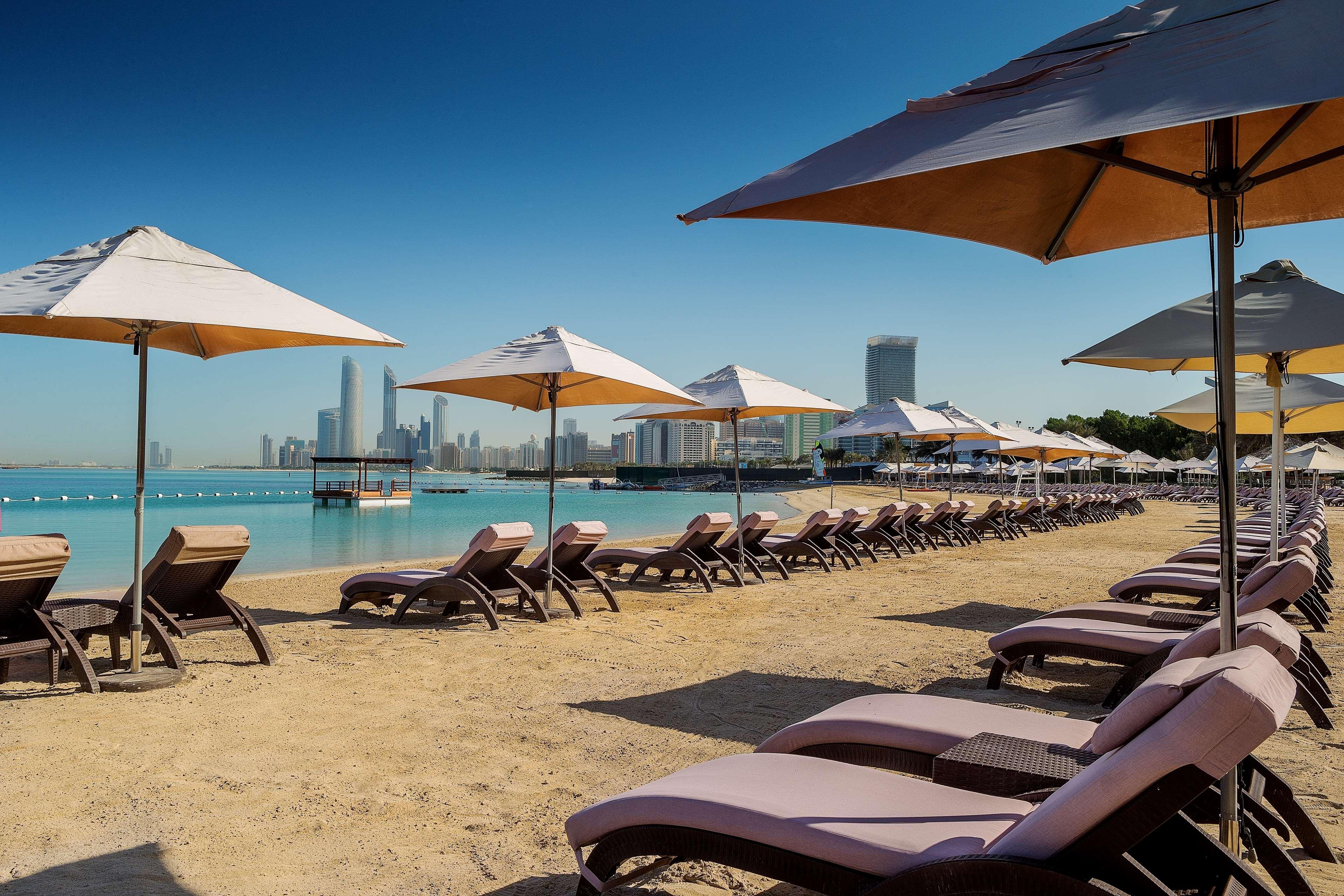 Radisson Blu Hotel & Resort, Abu Dhabi Corniche Esterno foto The photo depicts a serene beach scene featuring rows of lounge chairs and umbrellas arranged along the sandy shore. In the background, there are modern skyscrapers visible, suggesting a developed coastal city. The water is calm and turquoise, adding