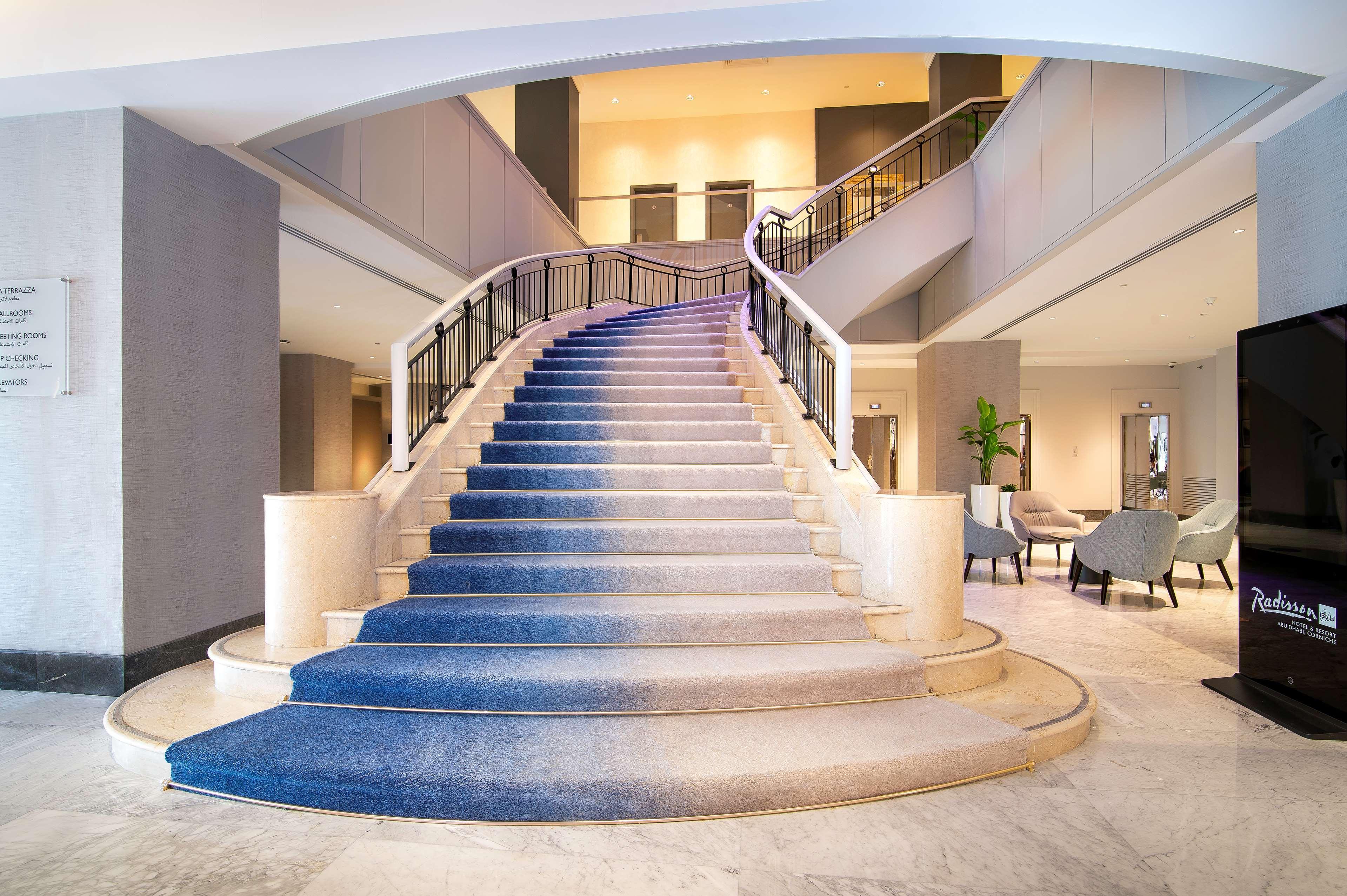 Radisson Blu Hotel & Resort, Abu Dhabi Corniche Esterno foto The photo depicts an elegant staircase inside a building, likely a hotel or a similar venue. The staircase is wide with a gentle curve, featuring a blue carpet that transitions in color from light to dark as it ascends. The walls surrounding the stai