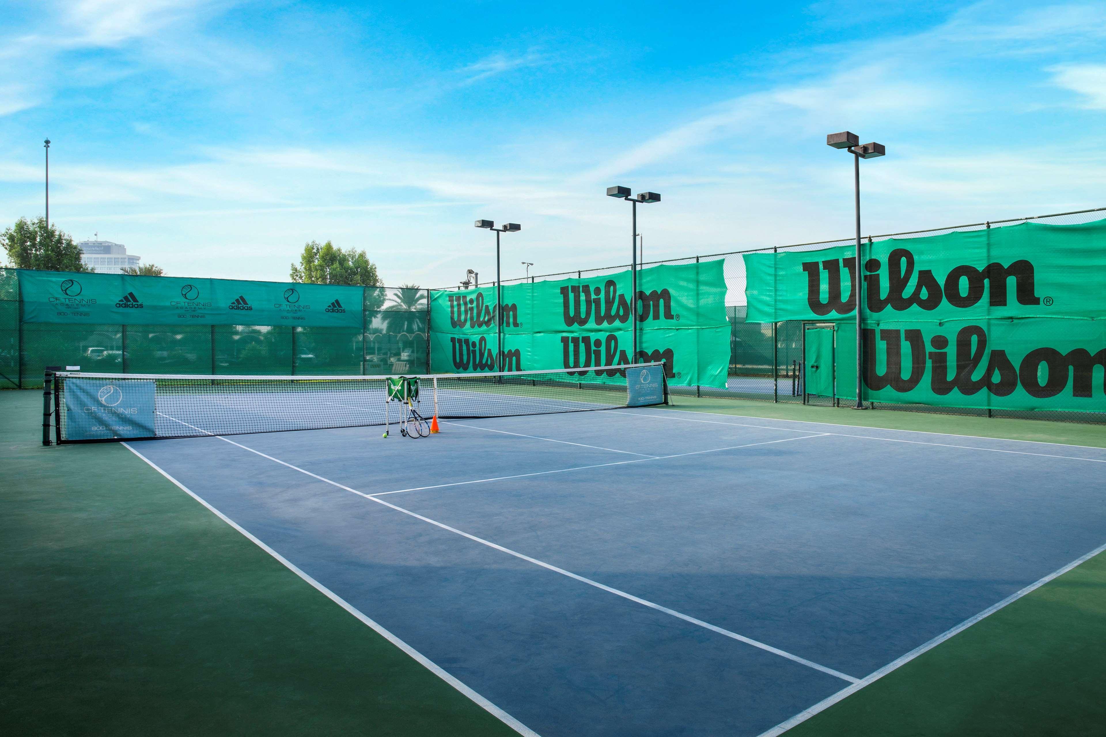 Radisson Blu Hotel & Resort, Abu Dhabi Corniche Esterno foto The photo shows a view of a tennis court surrounded by green nets and fencing featuring the brand name "Wilson." The court itself has a hard surface with marked lines for play. There are several tennis equipment items, such as a few rackets and possi