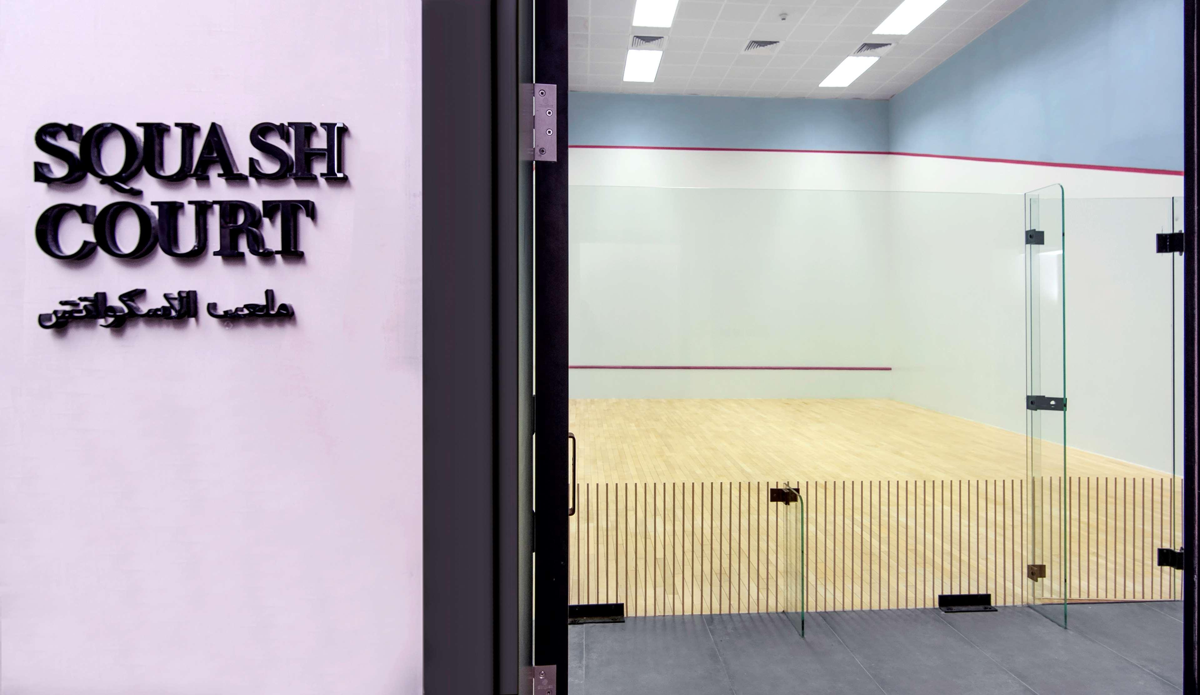 Radisson Blu Hotel & Resort, Abu Dhabi Corniche Esterno foto The photo shows the entrance to a squash court. There is a sign on the left side that reads "SQUASH COURT" in bold letters, with Arabic text beneath it. The court itself is visible through large glass doors, featuring a wooden floor and walls that ar