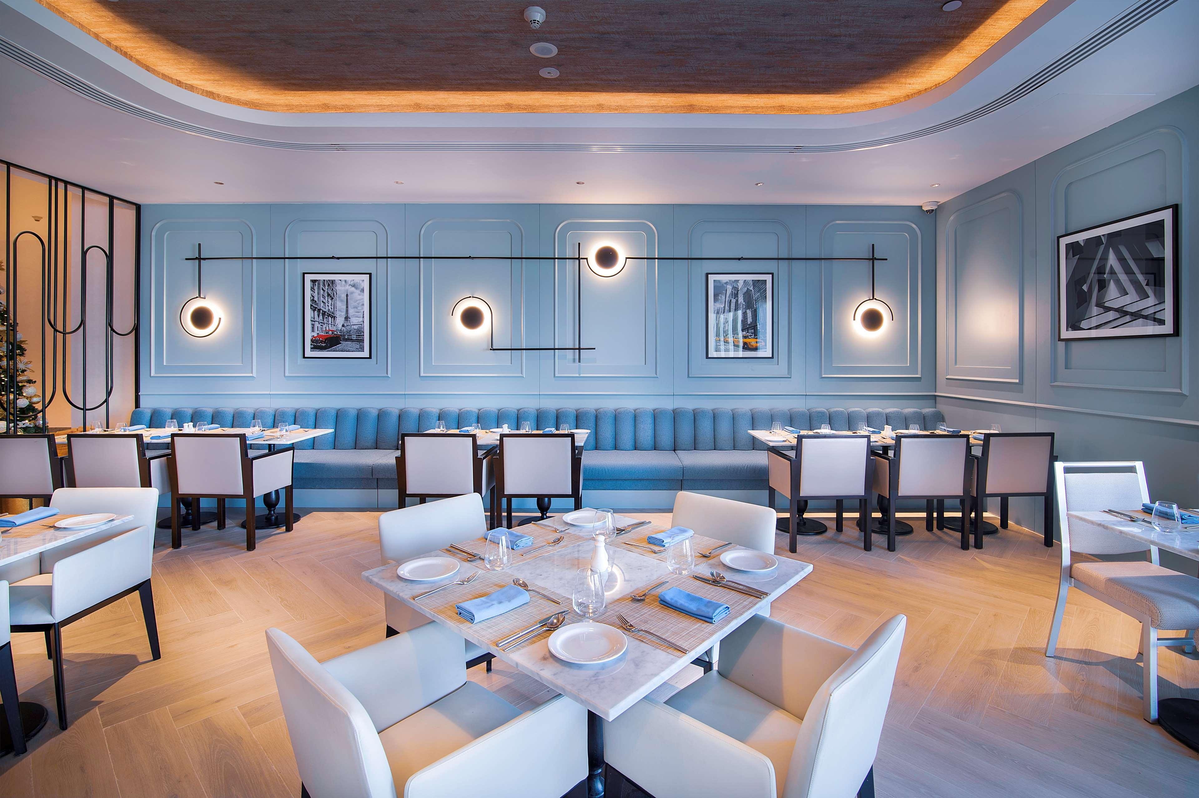 Radisson Blu Hotel & Resort, Abu Dhabi Corniche Esterno foto The photo shows a modern restaurant interior. The walls are painted in a soft blue, complemented by a decorative paneling design. There are multiple circular light fixtures hanging from the ceiling, providing ambient lighting. The seating includes a 