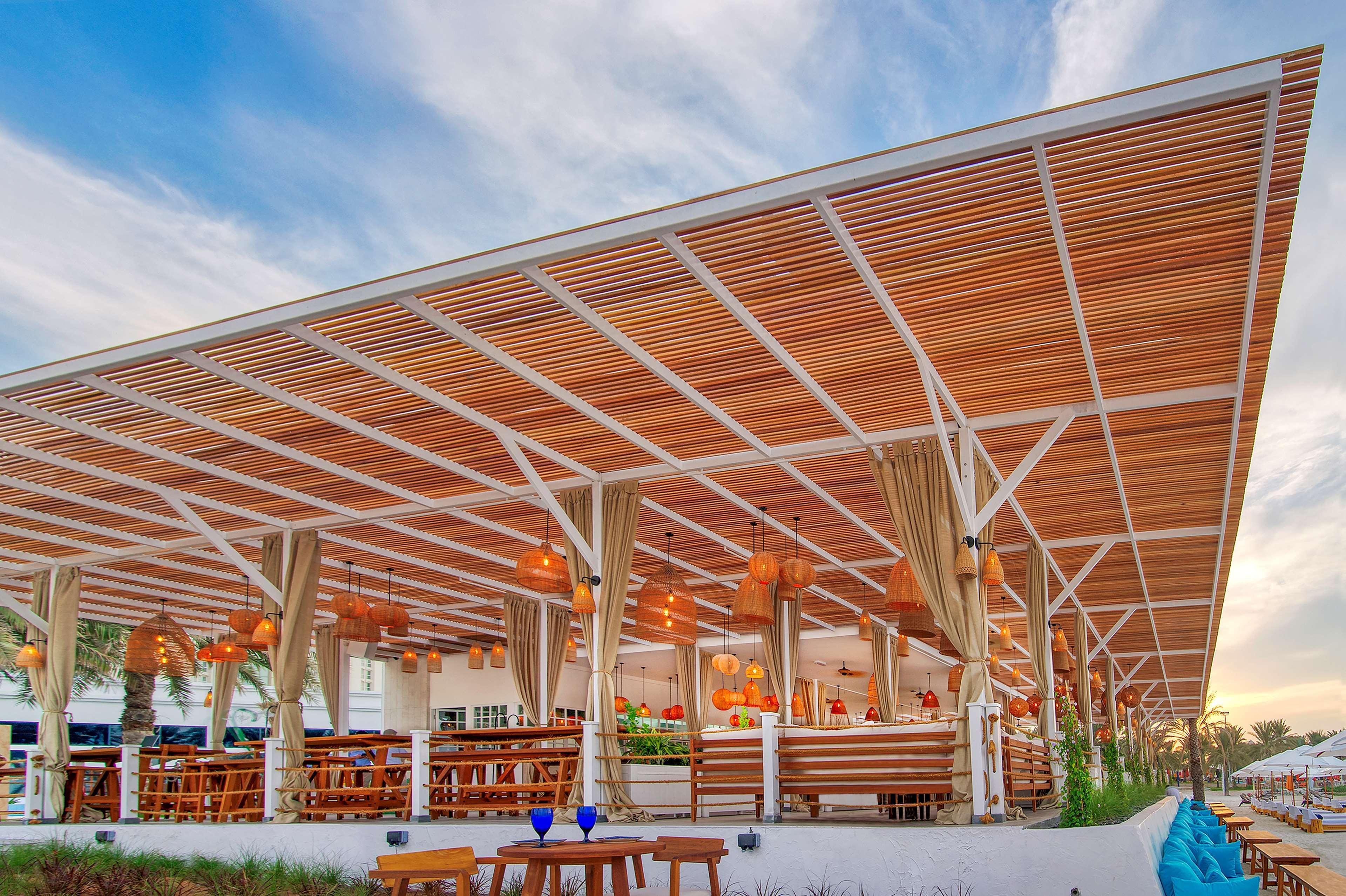 Radisson Blu Hotel & Resort, Abu Dhabi Corniche Esterno foto The photo shows a modern, open-air restaurant or café with a spacious wooden deck and a slatted roof. This design allows natural light to filter through, creating a bright ambiance. There are wooden furniture pieces, including tables and chairs, arra