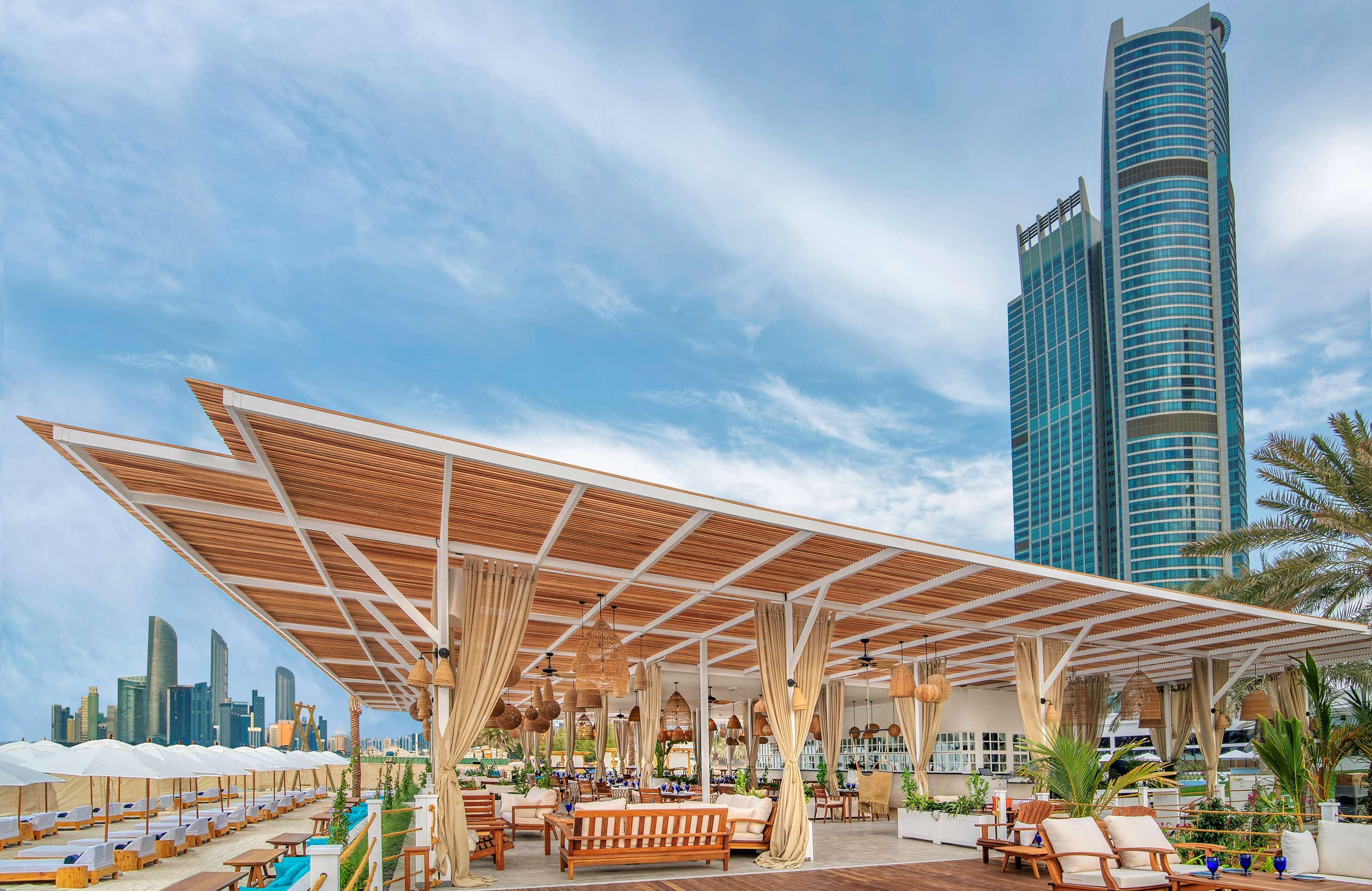 Radisson Blu Hotel & Resort, Abu Dhabi Corniche Esterno foto The photo shows a modern outdoor lounge or café area situated by a body of water, likely a beach or waterfront. The structure has a large, open roof with wooden beams and curtains providing shade. There are comfortable seating arrangements, such as s