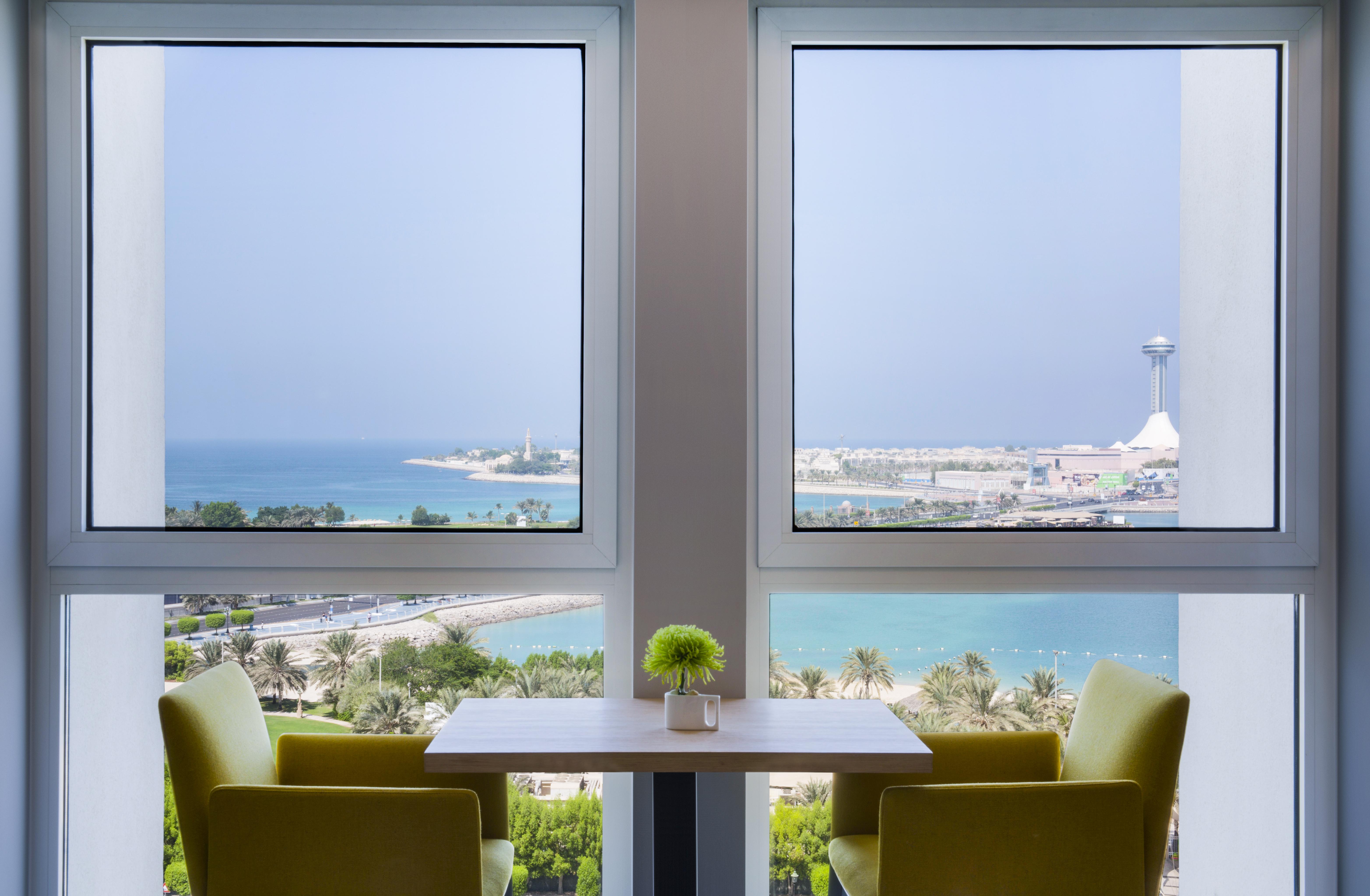 Radisson Blu Hotel & Resort, Abu Dhabi Corniche Esterno foto The photo shows a serene interior space featuring two large windows that frame a beautiful view of the ocean and coastline. The scene is bright, with sunlight filtering in, and there are two green chairs positioned around a small table. A small plant