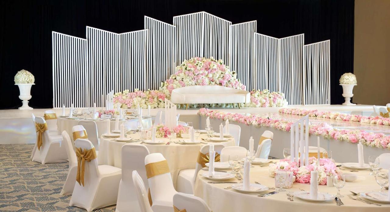 Radisson Blu Hotel & Resort, Abu Dhabi Corniche Esterno foto The photo features an elegantly decorated banquet hall set up for a special event, likely a wedding. In the background, there is a large backdrop adorned with vertical white lines and a lush arrangement of pink flowers. The seating area consists of r