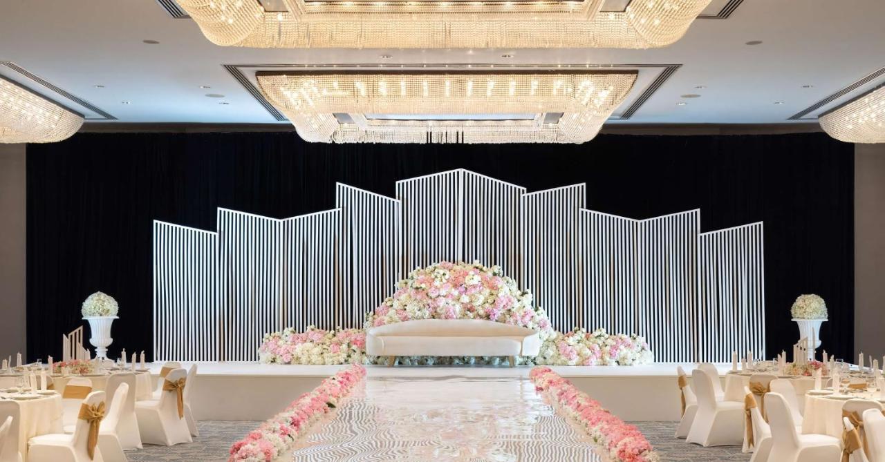 Radisson Blu Hotel & Resort, Abu Dhabi Corniche Esterno foto The photo shows a beautifully decorated wedding venue. In the center, there's a large, elegant white sofa on a stage, surrounded by a backdrop consisting of vertical white lines that create a modern, geometric design. In front of the sofa, a lush arr