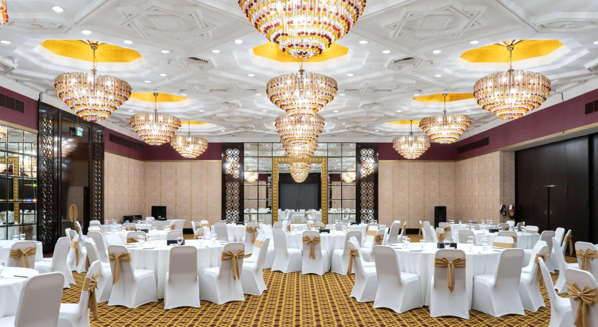 Radisson Blu Hotel & Resort, Abu Dhabi Corniche Esterno foto The photo depicts an elegant banquet hall or event space. The room is adorned with multiple large, ornate chandeliers that provide bright, ambient lighting. The tables are set with white tablecloths and chairs covered in white fabric, each decorated 
