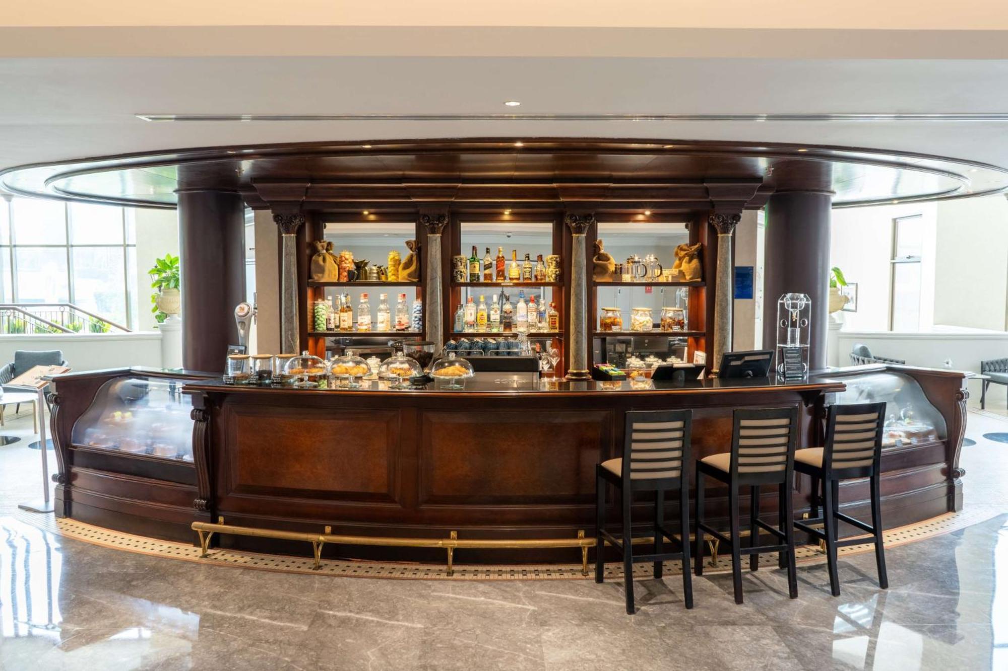 Radisson Blu Hotel & Resort, Abu Dhabi Corniche Esterno foto The photo shows a stylish bar area with a wooden bar counter. The bar has a polished surface and features a large wooden backlit shelving unit that holds various bottles of alcohol and glassware. There are decorative items placed on the shelves, addi